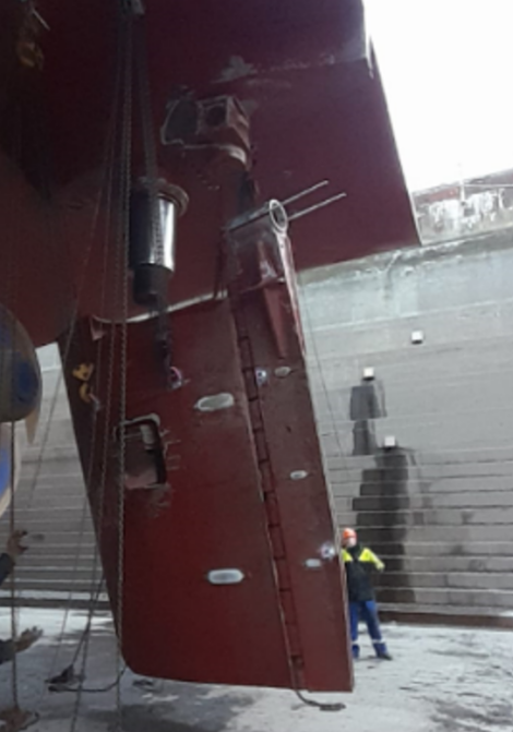 A worker stands next to a large ship that is in a dry dock, with the ship's rudder and propeller visible above the ground level.