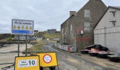 Road leading to mid yell harbour with speed limit signs and a dilapidated building with 'danger keep out' warnings.