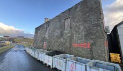 Weathered building with "danger" spray-painted on the side, flanked by construction containers under a cloudy sky.