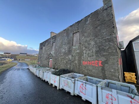 Weathered building with "danger" spray-painted on the side, flanked by construction containers under a cloudy sky.