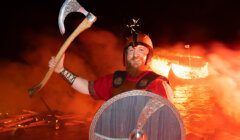 Person dressed as a viking holding an axe with a burning boat in the background.