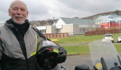 A smiling person holding a helmet stands next to a motorcycle in an outdoor setting with buildings in the background.