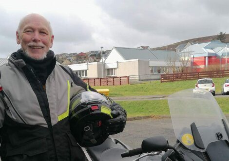 A smiling person holding a helmet stands next to a motorcycle in an outdoor setting with buildings in the background.