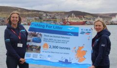 Two women standing outdoors, holding a sign that celebrates scottish fishermen for removing 2,300 tonnes of litter from the ocean.