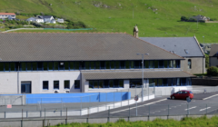 Modern school building with a blue fence in a green hilly landscape.