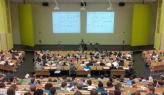 Lecture in a university auditorium with students watching a presentation involving mathematical equations.