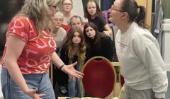 Two women engaged in a heated conversation with a group of onlookers showing varied reactions in the background.