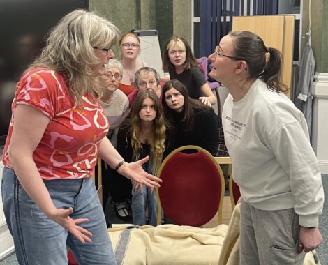 Two women engaged in a heated conversation with a group of onlookers showing varied reactions in the background.