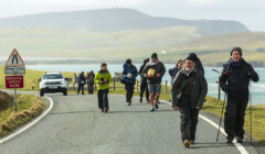 Group of hikers walking along a single-track road near a coastal area with a warning sign advising to reduce speed now.