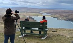 A person capturing a photograph of two individuals sitting on a bench overlooking a scenic coastal landscape.