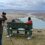 A person capturing a photograph of two individuals sitting on a bench overlooking a scenic coastal landscape.