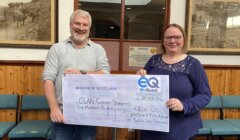 Two individuals holding a large charity check from bank of scotland inside a room with historical photos on the wall.