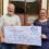 Two individuals holding a large charity check from bank of scotland inside a room with historical photos on the wall.