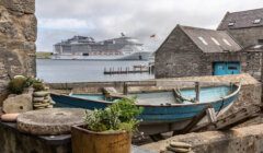 Old wooden boat and stacked stones in the foreground with a large cruise ship anchored in the bay beyond, viewed from a coastal village.