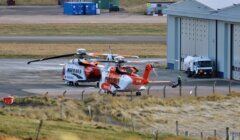 Two helicopters on a tarmac near an airport building with ground support equipment in the vicinity.