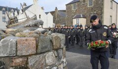 A uniformed individual laying a bouquet of flowers with a group of solemn onlookers in the background.