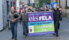 A group of demonstrators marching on a street, led by a bagpiper, while carrying a banner for the further education lecturers' association advocating for unity and strength.