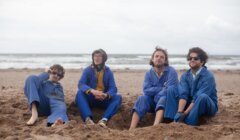 Four men in coveralls sitting on a sandy beach.