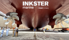 A large ship in dry dock with workers standing below its massive hull and propellers.