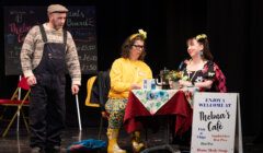 Three actors on stage in a scene set in a cafe, with one standing and two seated at a table, engaging in conversation amidst props and a menu sign.