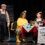 Three actors on stage in a scene set in a cafe, with one standing and two seated at a table, engaging in conversation amidst props and a menu sign.