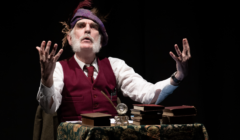 A person performing on stage, dressed in vintage attire with books and a pocket watch on a table, portraying a dramatic expression with raised hands.