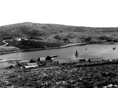 Rural coastal village with scattered buildings and a boat on the water, surrounded by rolling hills.