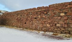 A weathered stone wall on a snow-dusted ground with patches of grass.