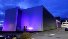 A large modern building illuminated with purple lights under an evening sky, with a parking lot in the foreground.