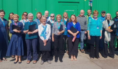 Group of smiling adults in casual and professional attire with lanyards standing in front of a green wall under sunlight.