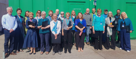 Group of smiling adults in casual and professional attire with lanyards standing in front of a green wall under sunlight.