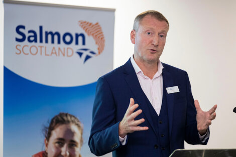 A man speaking at a podium with a "salmon scotland" banner in the background; he gestures with his hands as he talks.