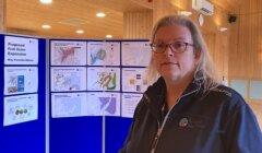 Woman in uniform standing in front of informational posters about a proposed fish holm expansion.