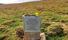 A memorial stone with an inscription and flowers on top sits in a grassy field under a cloudy sky.