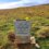 A memorial stone with an inscription and flowers on top sits in a grassy field under a cloudy sky.