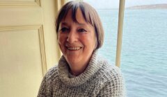 A smiling middle-aged woman in a gray knitted sweater standing next to a window with a view of the sea.