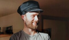 A young man with a beard, wearing a flat cap, smiles gently in a warmly lit room.