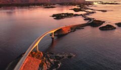 Aerial view of a curvy bridge crossing over a series of small islands in a calm sea at sunset, with mountains in the background.