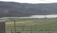 A scenic view of a rural landscape featuring a lake surrounded by rolling hills, with a line of power poles and cables in the foreground.