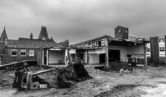 Black and white photo of a dilapidated building with a collapsed roof and scattered debris, featuring a toppled excavator in the foreground.