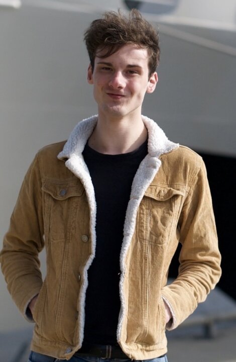 Young man in a tan corduroy jacket with white lining, smiling, standing outdoors with a blurred background.