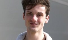 Young man with a slight smile, wearing a brown jacket with a white fleece collar, standing outdoors.