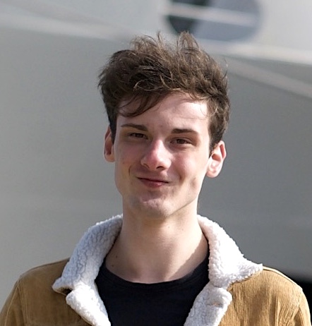 Young man with a slight smile, wearing a brown jacket with a white fleece collar, standing outdoors.
