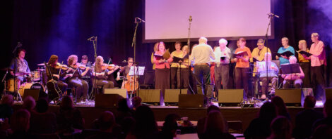 A live concert scene with a choir and orchestra performing on stage in front of an audience under purple stage lighting.