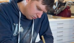 A focused young man in a hoodie drawing carefully with a pencil on a sheet of paper at a desk indoors.