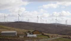 A rural landscape featuring a small road, two houses, and a large wind farm with multiple turbines on a hilly terrain under a cloudy sky.