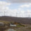 A rural landscape featuring a small road, two houses, and a large wind farm with multiple turbines on a hilly terrain under a cloudy sky.