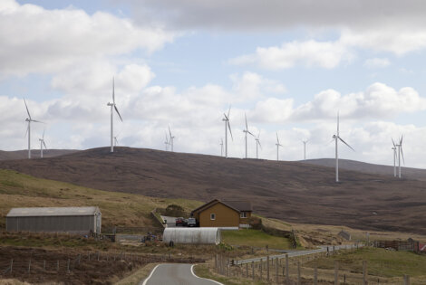 A rural landscape featuring a small road, two houses, and a large wind farm with multiple turbines on a hilly terrain under a cloudy sky.