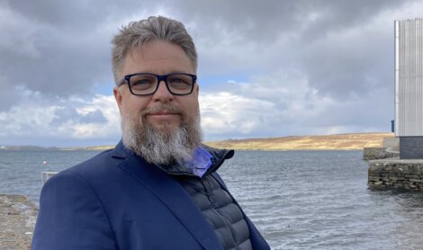 A man with glasses and gray hair stands by a lake with hills in the background, wearing a blue jacket and vest.