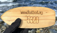 A hand wearing a glove holds an oval wooden tag engraved with "www.floatboat.org 5320" against a backdrop of a rippling body of water.